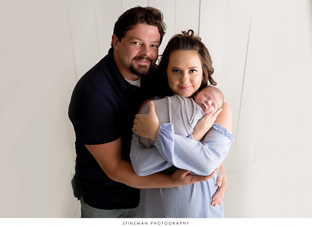 Mom and dad holding their newborn baby boy in newborn photo shoot in south jersey. Baby is wearing grey, mom is wearing baby blue and dad is wearing a navy blue polo for their session. 
