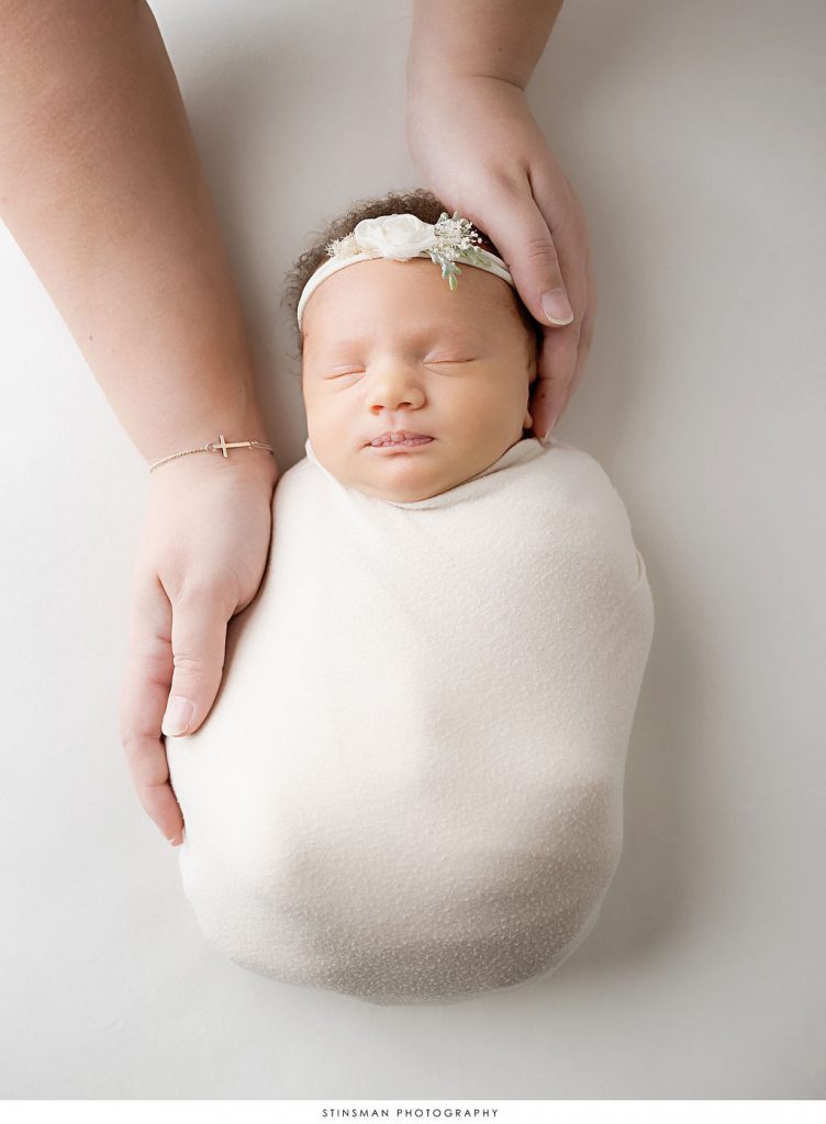 Newborn baby girl asleep with mother's hands in a white swaddle with a floral headband during newborn photoshoot.