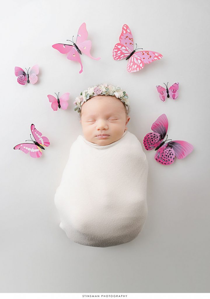 Newborn baby girl swaddled in white surrounded by butterflies during her newborn photoshoot