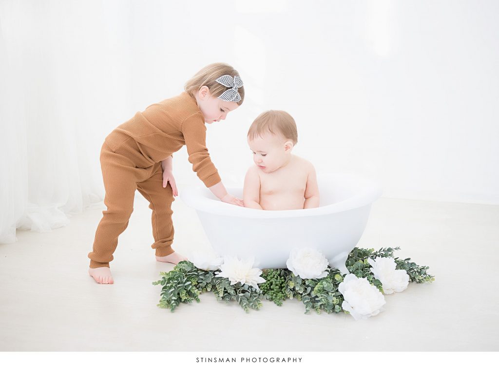 Big sister showing little sister how to play in the tub at her first birthday milestone photoshoot