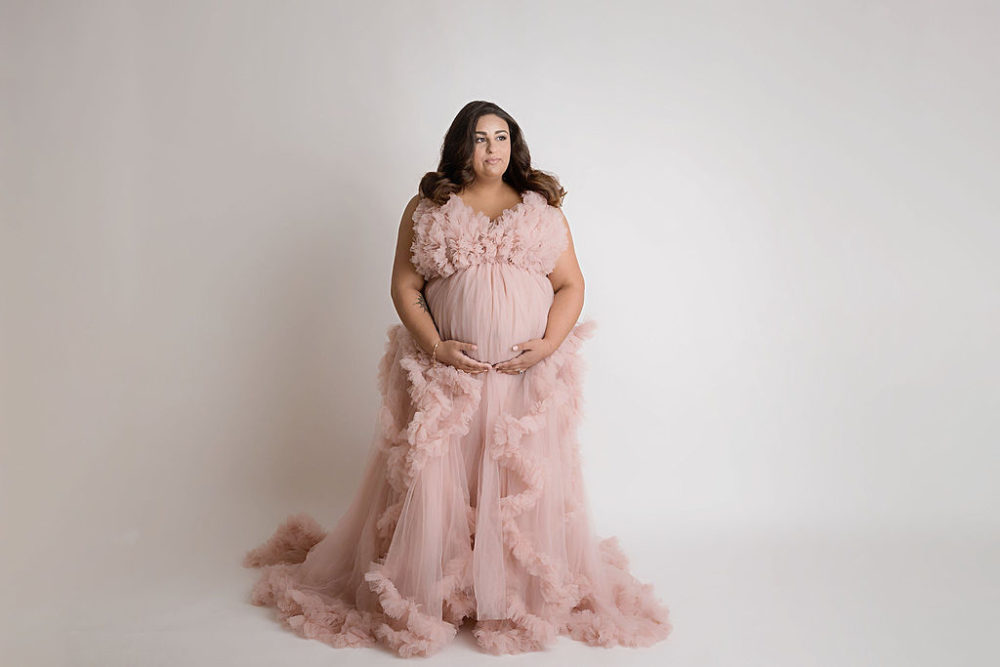 Woman holding belly wearing ruffle dress for her pop of pink in-studio maternity session in South Philadelphia