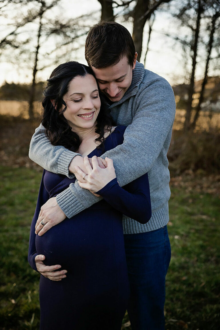 Man and woman embracing each other while holding belly for navy blue maternity session in Southampton, New Jersey.