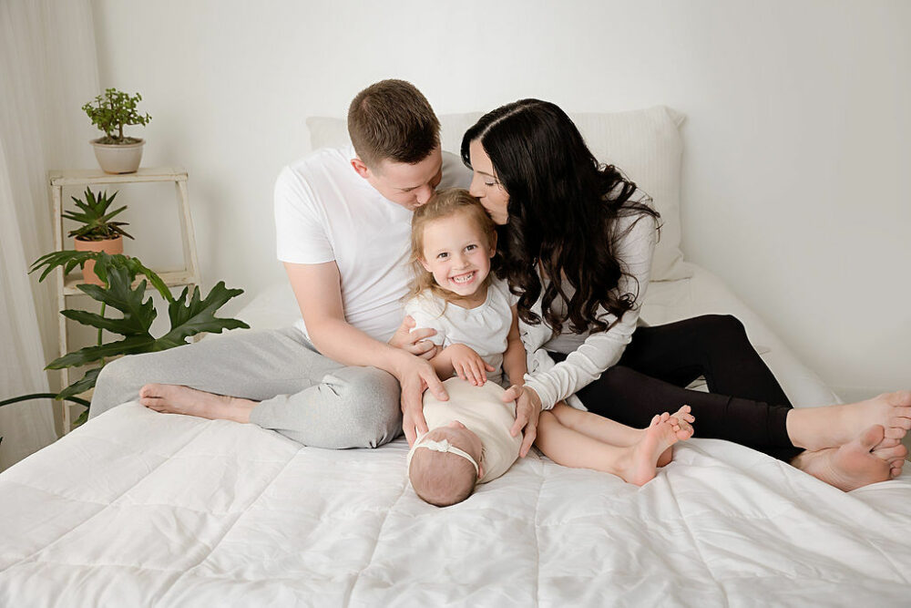 Parents kissing big sister while holding newborn baby for newborn session in Clifton, New Jersey.
