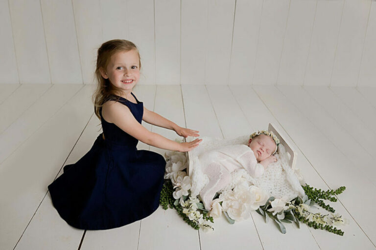 Big sister sitting and holding baby photography props where baby sister is sleeping for a navy blue maternity and newborn session in Cape May, New Jersey.