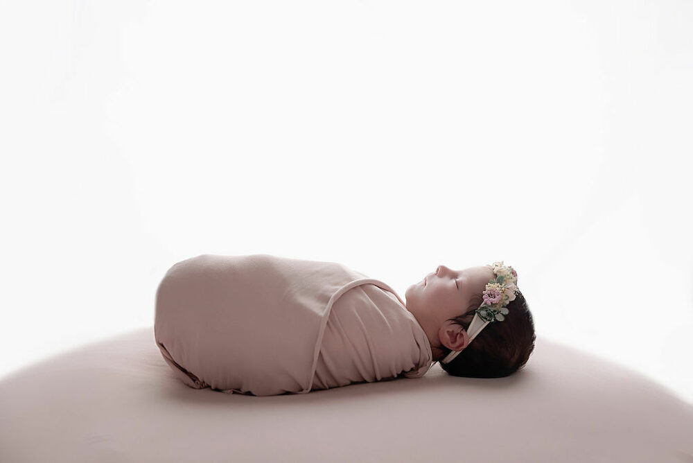 Sleepy newborn girl wrapped in swaddle wearing headband for her in studio professional baby photos taken in Glassboro, New Jersey.