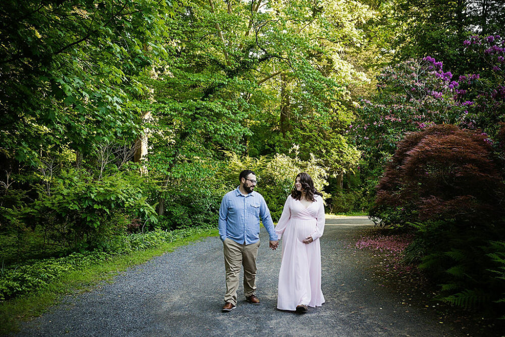 Parents to be holding hands and walking through garden for their couple maternity photos in Hamilton, New Jersey.