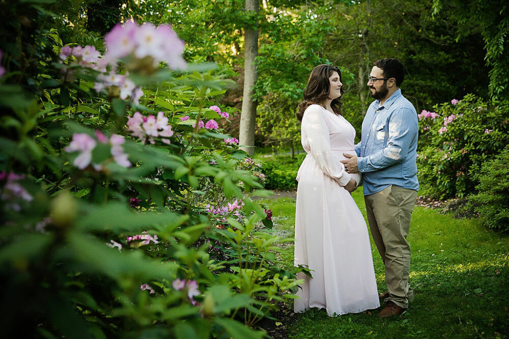 Husband and wife holding her pregnant belly smiling at each other for their pregnancy pictures taken during her Spring garden maternity session in Burlington, New Jersey.