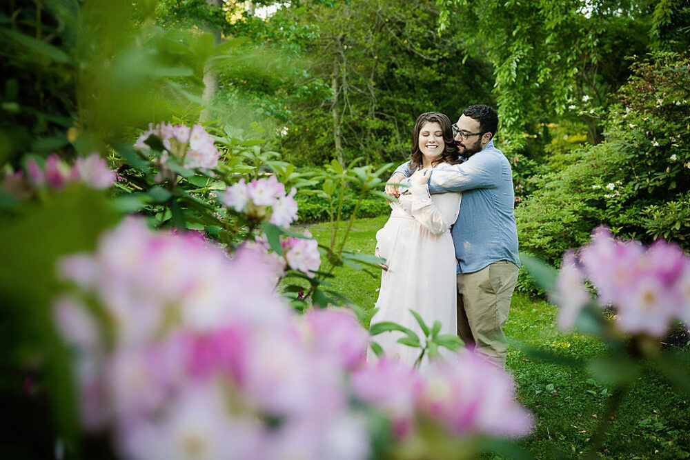 Husband hugging pregnant wife while she's holding belly for their unique maternity pictures for her spring garden maternity session in Haddonfield, New Jersey.