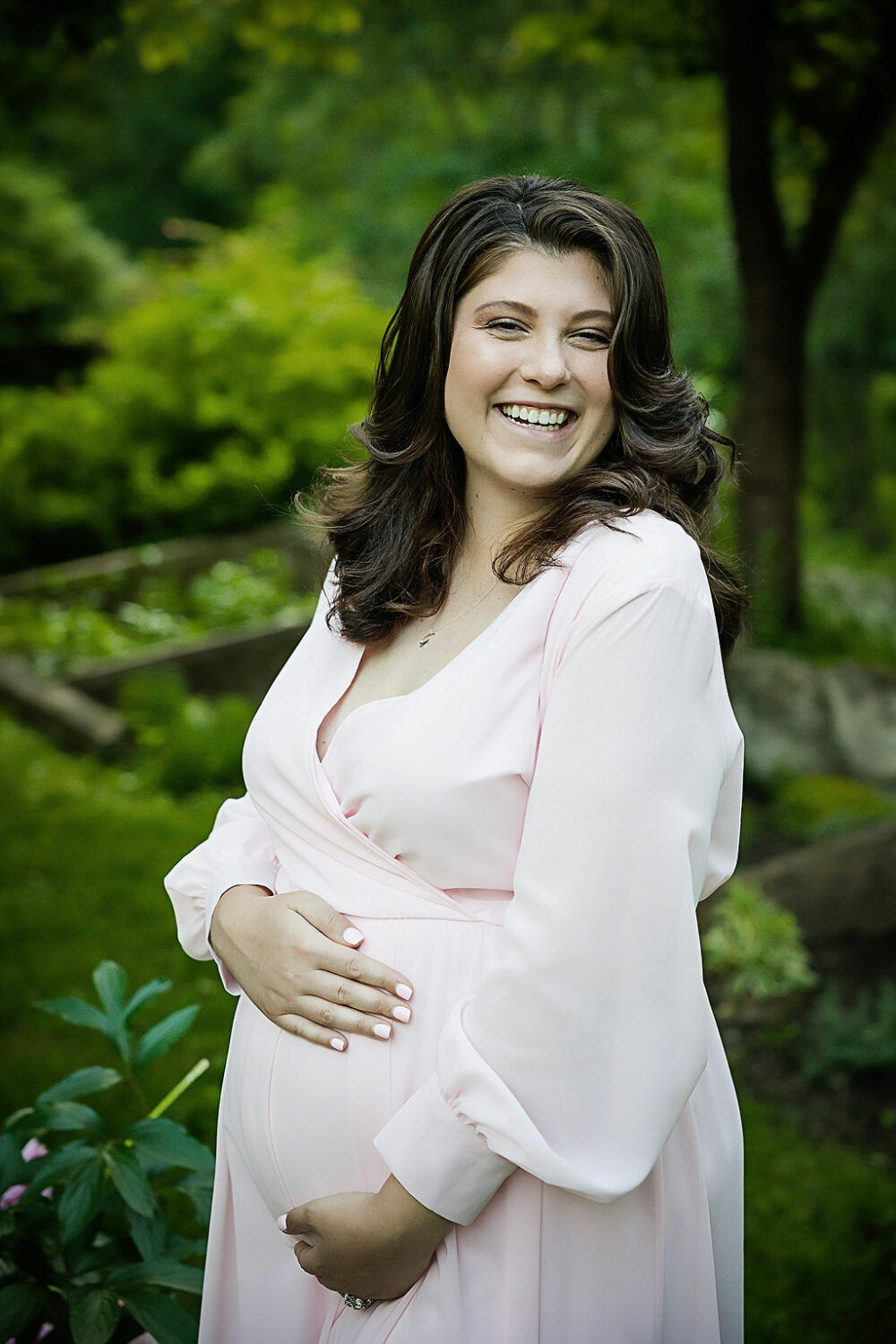 Pregnant woman holding belly wearing long sleeve maternity dress doing cute maternity poses for her spring garden maternity session at Sayen Gardens, New Jersey.