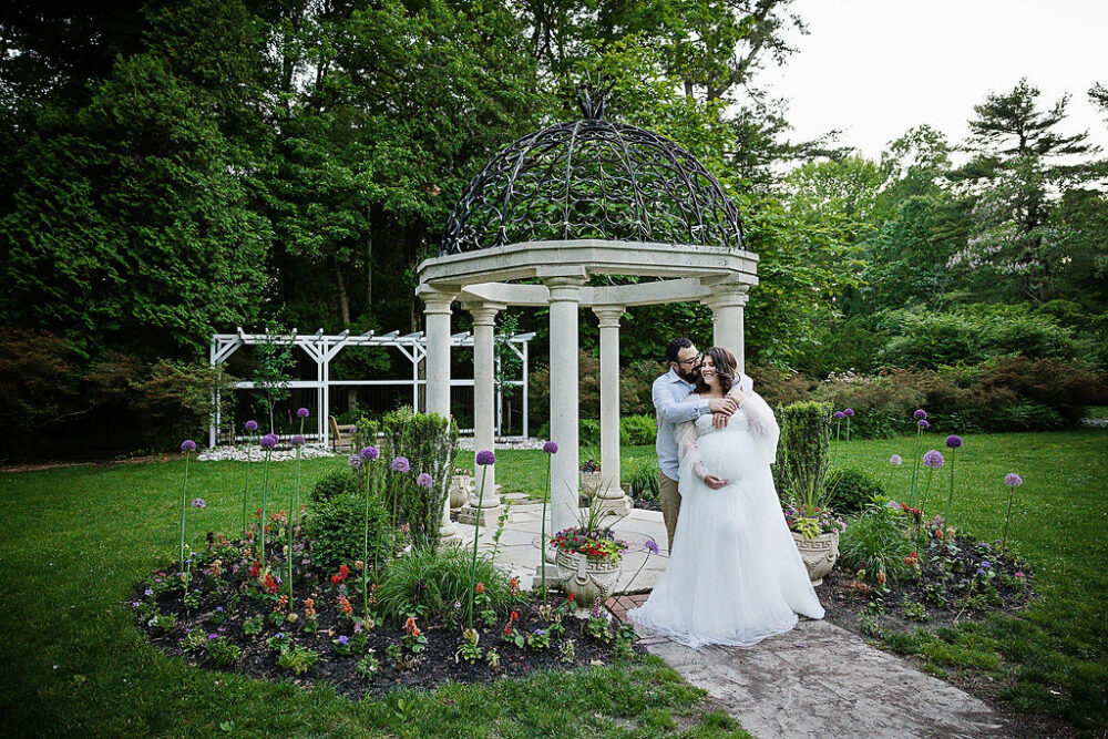 Husband embracing why is why she holds her pregnant belly standing near gazebo for their Spring Garden maternity session in Barrington, New Jersey.