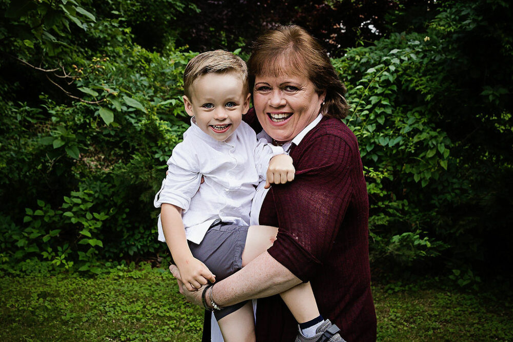 Grandmother holding her grandson in arms and smiling at camera with green landscape background for their estate lifestyle family session taken in Camden, New Jersey