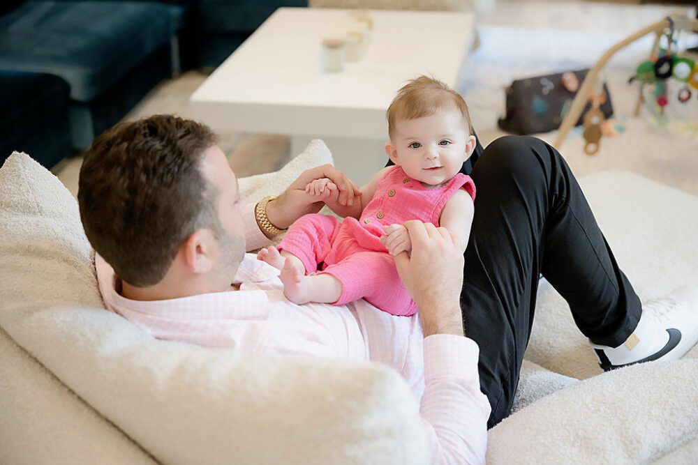 Three month old girl sitting on her father's stomach in their living room for their lifestyle family session taken in Mount Holly, New Jersey.