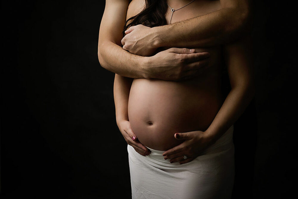 Close-up picture of pregnant woman holding pregnant belly and husband embracing her from behind for their nude in-studio maternity session in Camden, New Jersey.