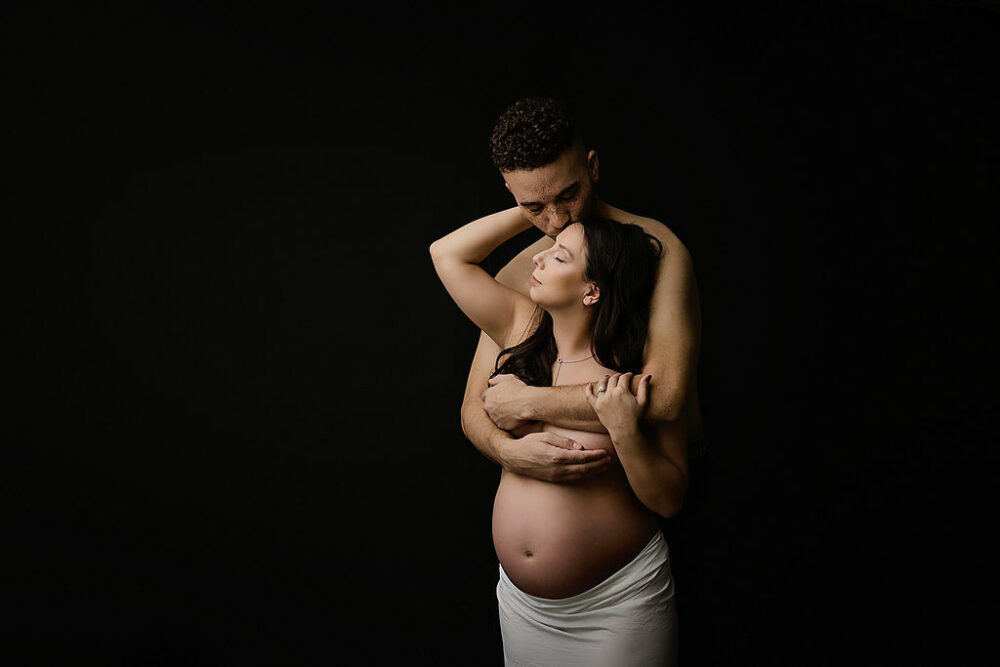 Intimate photo of husband, kissing wife on forehead while embracing each other against a black backdrop for their shirtless nude in-studio maternity session in Burlington, New Jersey.