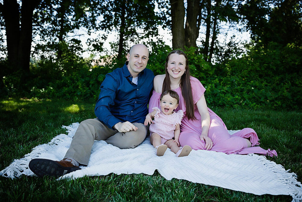 Parents and young daughter sitting on picnic blanket outdoors for their daughter’s pink first birthday session in collingswood, New Jersey.