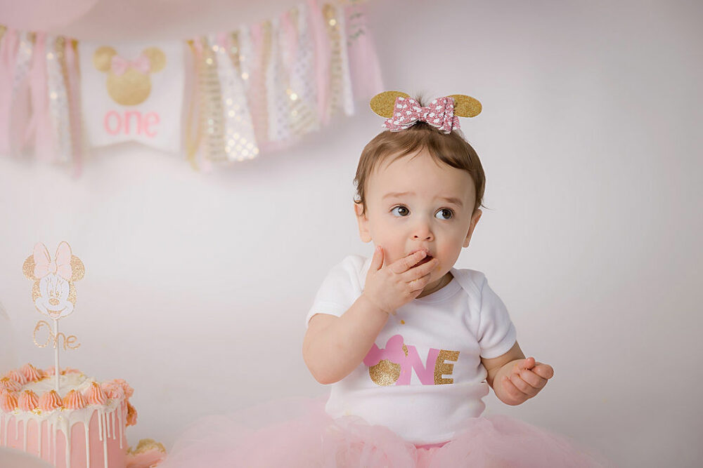 Toddler girl looking up wearing Minnie mouse, bow and shirt for her pink baby cake smash photos taken in Bellmawr, New Jersey.