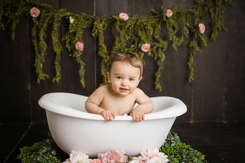 Toddler girl, smiling at camera and baby bathtub prop for he pink first birthday session in Southampton, New Jersey.