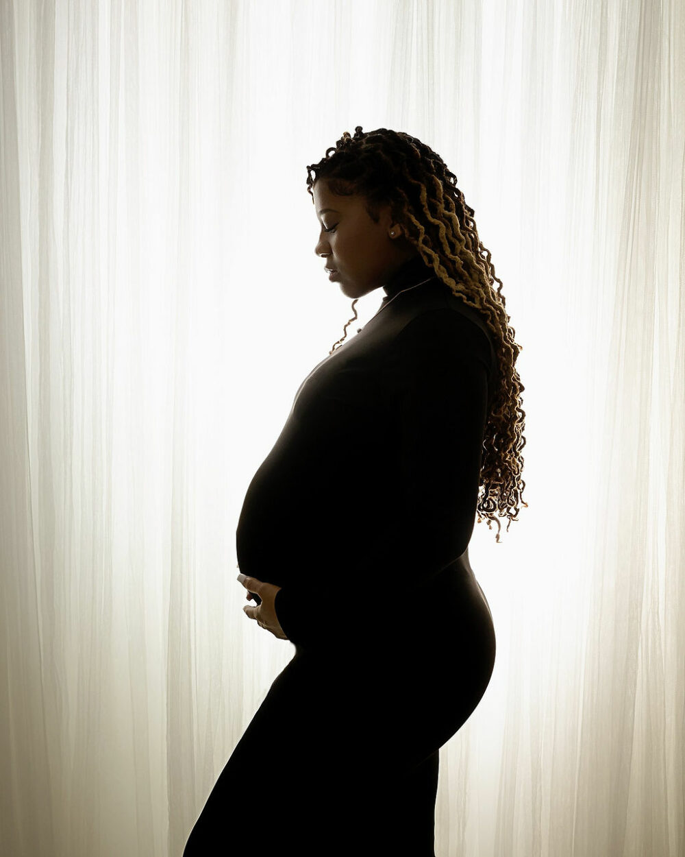 Side profile portrait of Woman wearing black body con dress against light backlit backdrop for her angelic maternity session taken in freehold, New Jersey.
