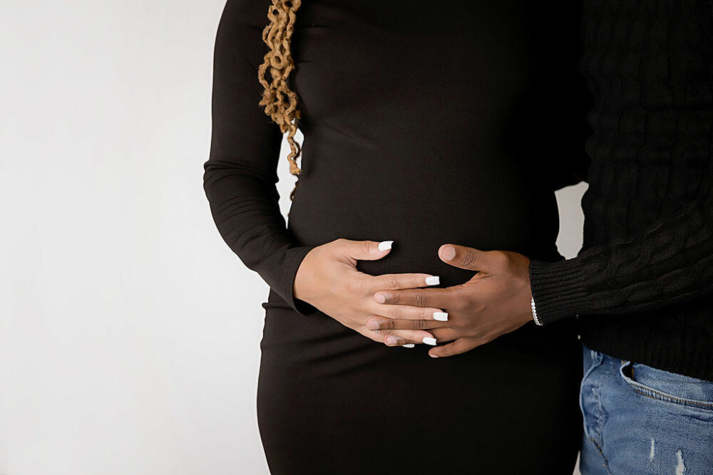 Close up of couple maternity photo of woman and man holding baby bump with fingers intertwined for their black maternity pictures taken in Millville, New Jersey.