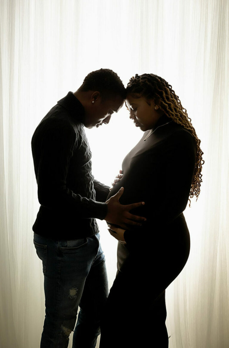 Pregnant woman and boyfriend holding baby bump and touching foreheads against a bright backdrop for their couple maternity photos taken in Cinnaminson, New Jersey.