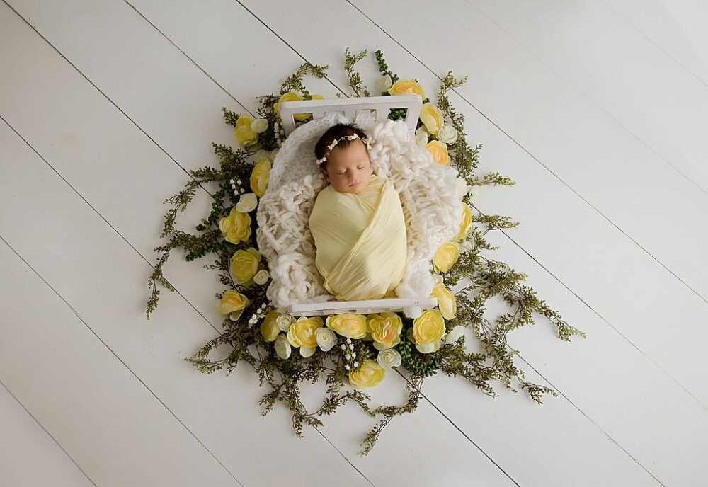 Sleepy newborn girl and swaddle on newborn photography props for her yellow in studio newborn session in Southampton, New Jersey.