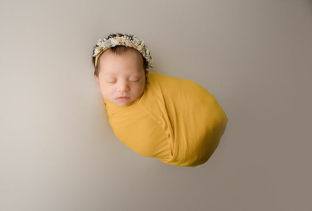 Newborn girl wrapping in muslin and wearing headband for her baby photoshoot ideas for her yellow in-studio newborn session in Camden, New Jersey.