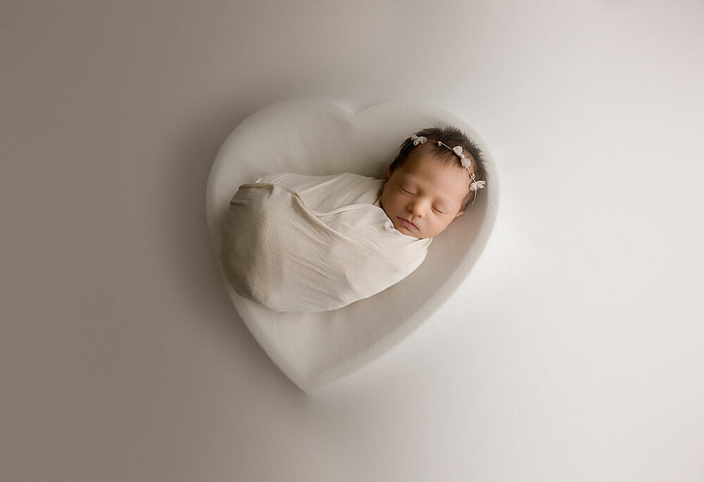 Newborn girl in swaddle, sleeping on heart, photography prop for her in-studio baby photography In Cherry Hill, New Jersey.
