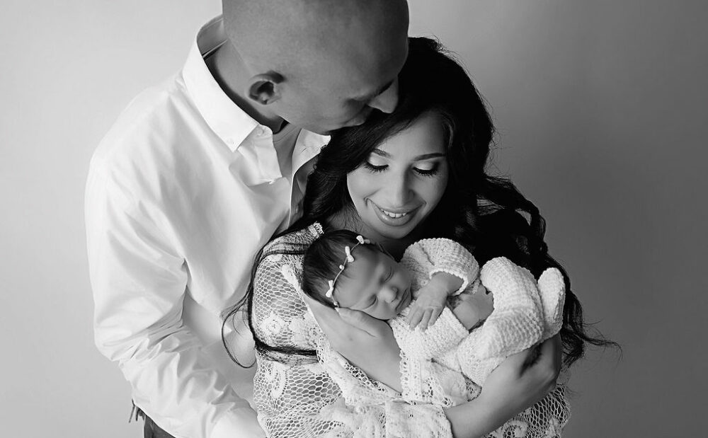 Black and white portrait of husband, kissing wife while she’s holding newborn girl for their yellow in studio newborn session, taken in Pemberton, New Jersey.