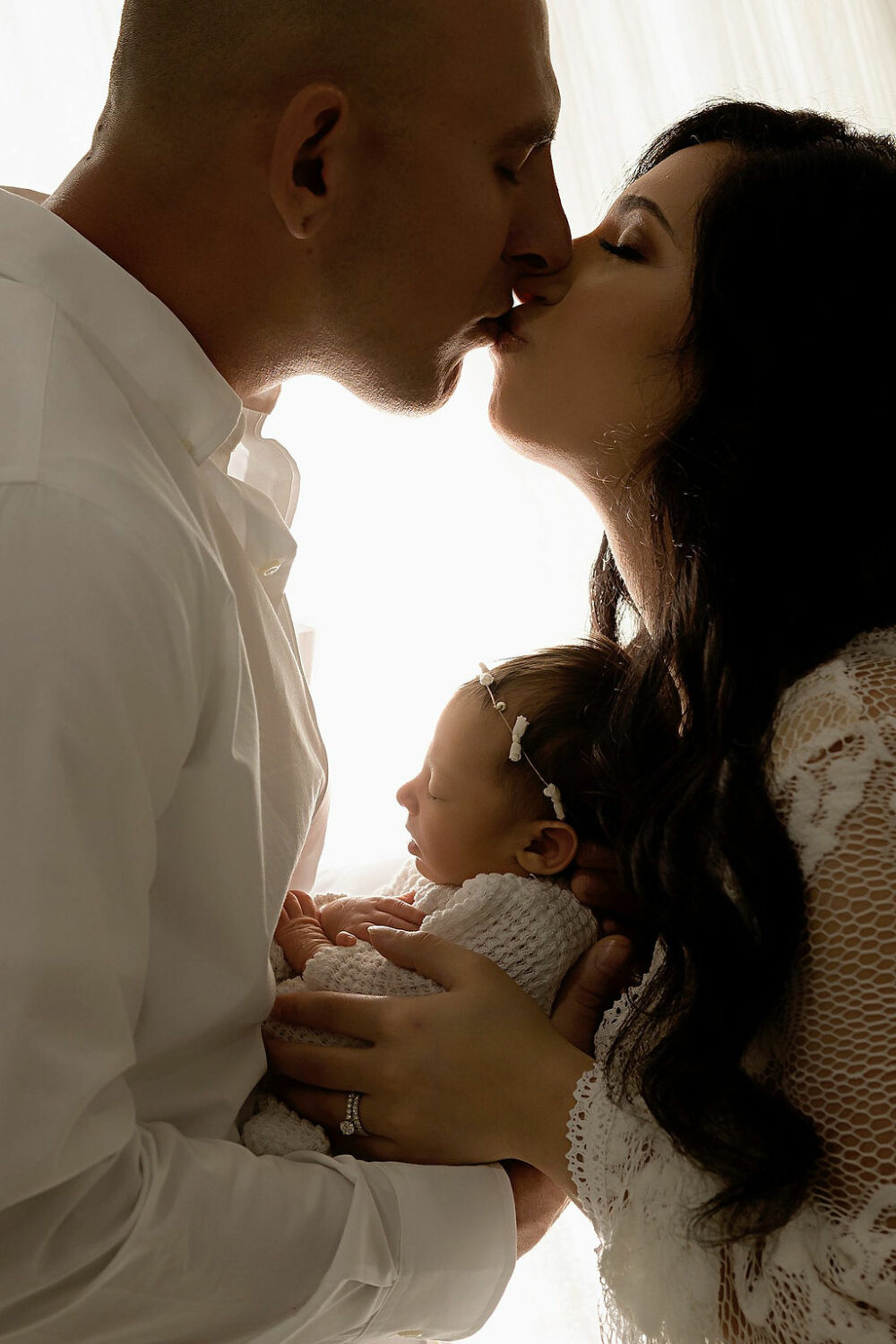 Close up back lit image of husband and wife, kissing while holding newborn and their arms for their newborn family pictures taken in studio in Moorestown, New Jersey.