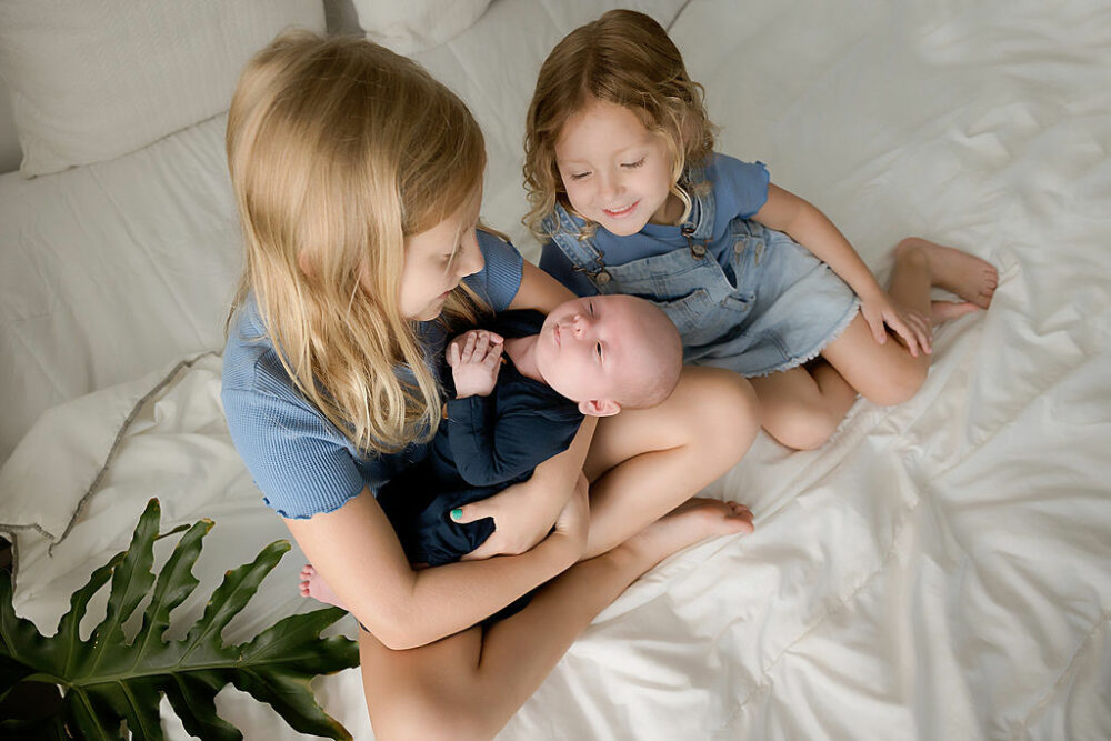 Two sisters, smiling, and looking at newborn brother, sitting on bed, photography prop for their blue newborn session taken in studio in Monmouth County, New Jersey.