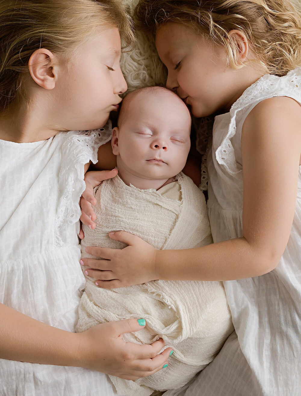 Close-up of two sisters, holding newborn, sleeping brother in between them, laying down and kissing his head for their baby photography session in holmdel, New Jersey.