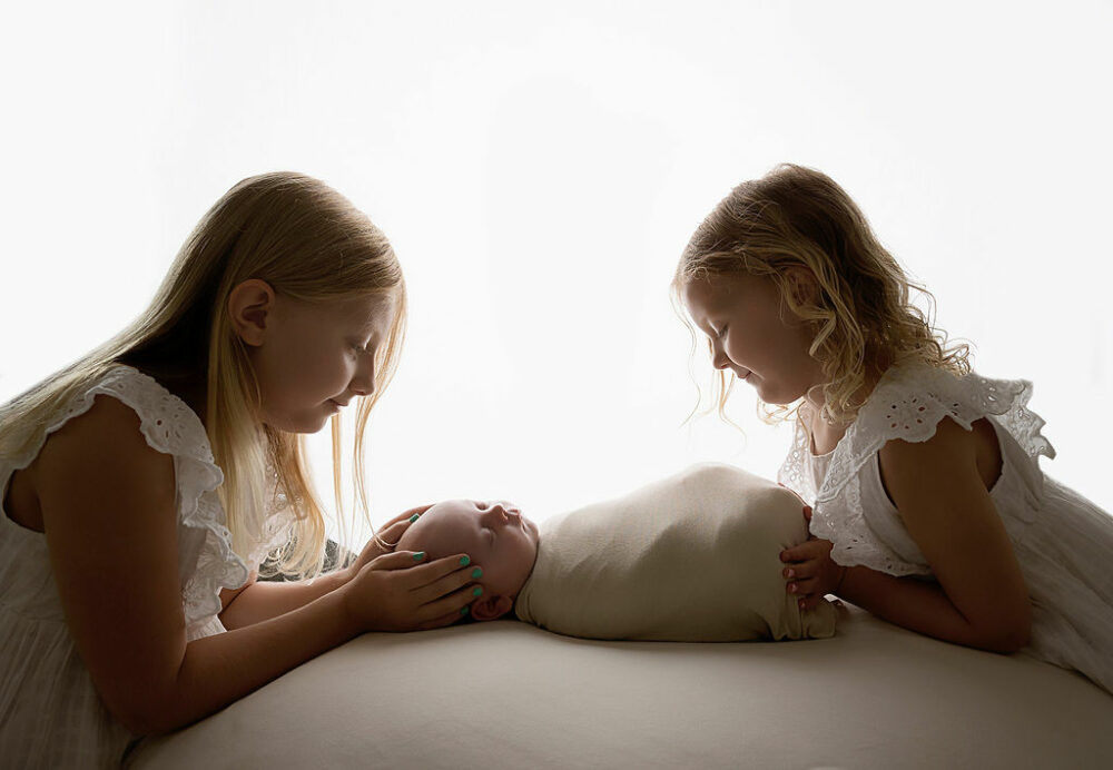 Light and bright sibling portrait of two sisters, holding their sleepy newborn brother against a white light background for baby photo, shoot ideas taken in studio in Millstone, New Jersey.