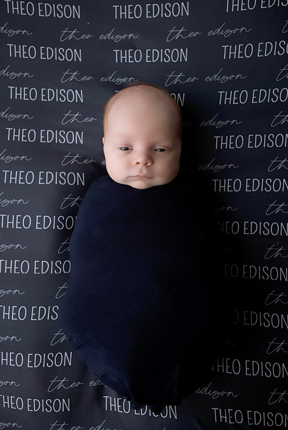 Infant boy, swaddled on personalized blanket, awake for his blue newborn photography session taken in Camden, New Jersey.