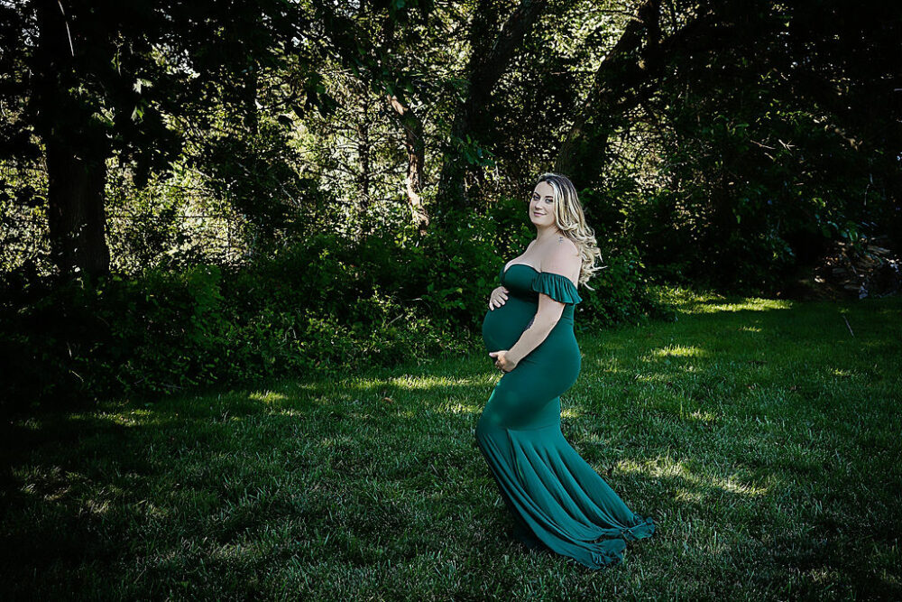 Pregnant women holding her belly while wearing green maternity dress, and smiling at the camera posing outside for her maternity photo, shoot ideas taken in Moorestown, New Jersey.