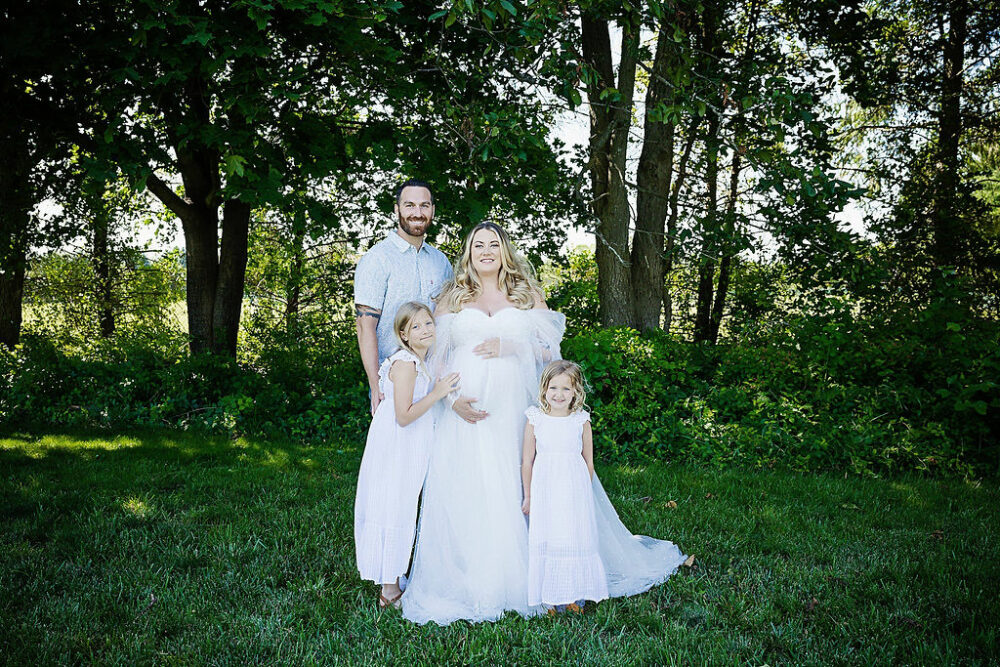 Pregnant women holding belly, wearing scalloped white dress, standing next to her husband, and two daughters, while holding her belly for professional maternity pictures, taken in Warren, New Jersey.