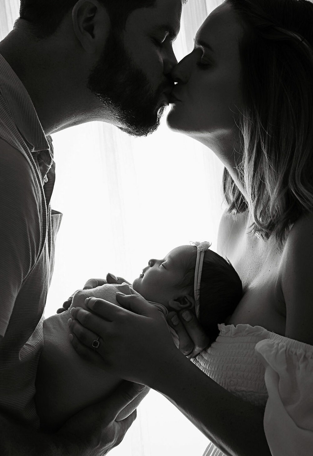 Close up black and white photo of husband and wife kissing against light and bright backdrop while holding newborn girl for their newborn session taken in Mount Holly, New Jersey.
