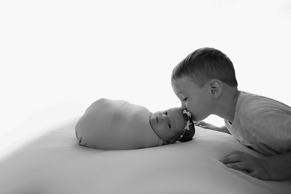 Black and white sibling portrait of toddler boy kissing his newborn sister on head while she's lying awake for her in-studio lavender newborn session in Millburn, New Jersey.