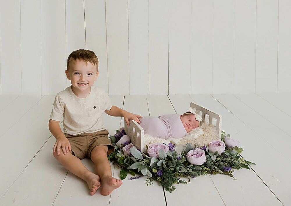 Toddler boy sitting next to sleeping baby sister smiling a camera for there in studio lavender newborn session taken in chatham, New Jersey.