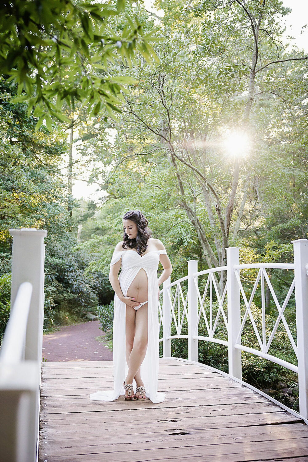 Pregnant woman on wooden bridge and garden holding her belly with her legs crossed and maternity dress for her mile stone maternity and newborn session in Mount Holly, New Jersey.