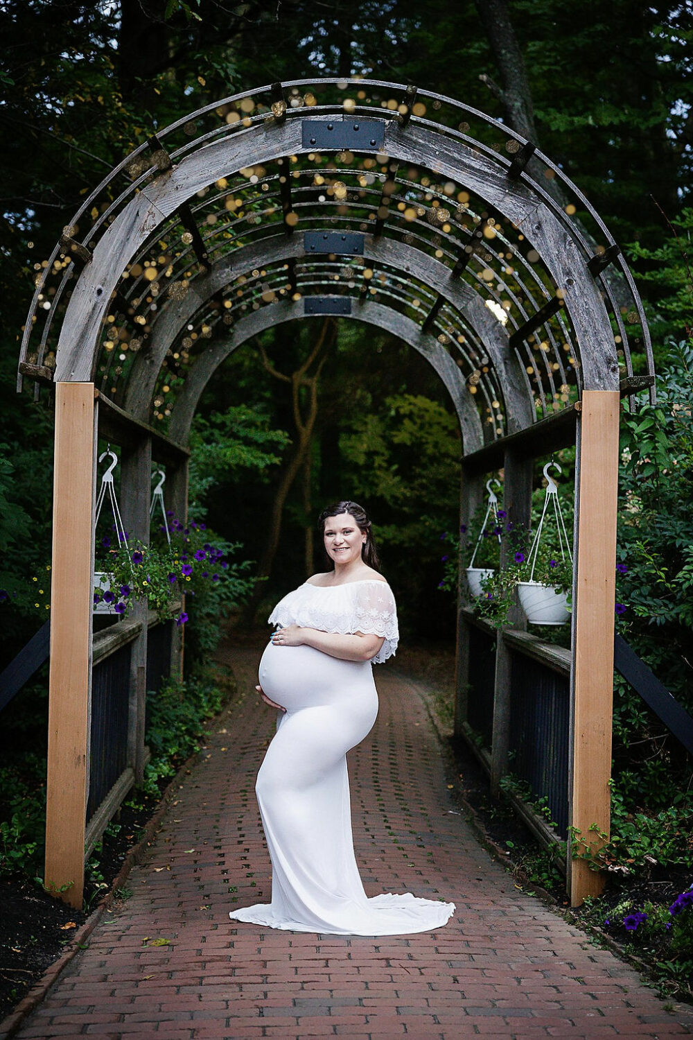 Woman smiling at camera holding belly wearing dress on garden trail for her maternity picture ideas taken in Burlington, New Jersey.