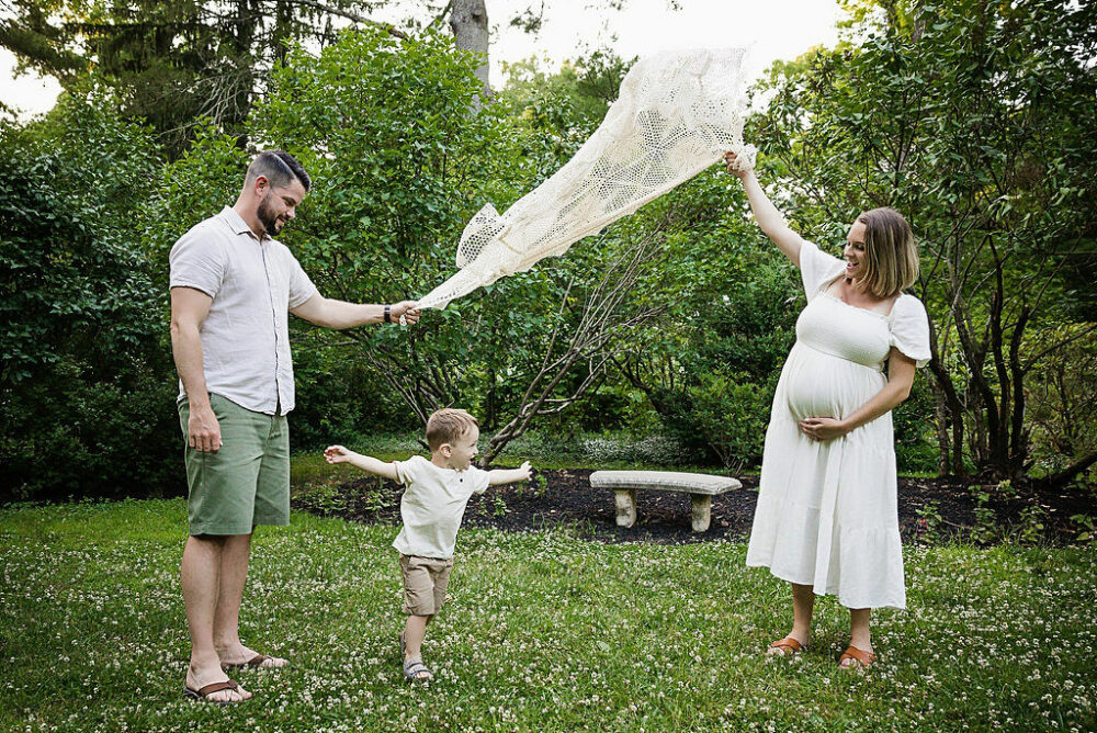 Lifestyle photo of husband and wife playing in the garden with their toddler boy while mother is holding her pregnant belly for their lavender maternity session in Ridgewood, New Jersey.
