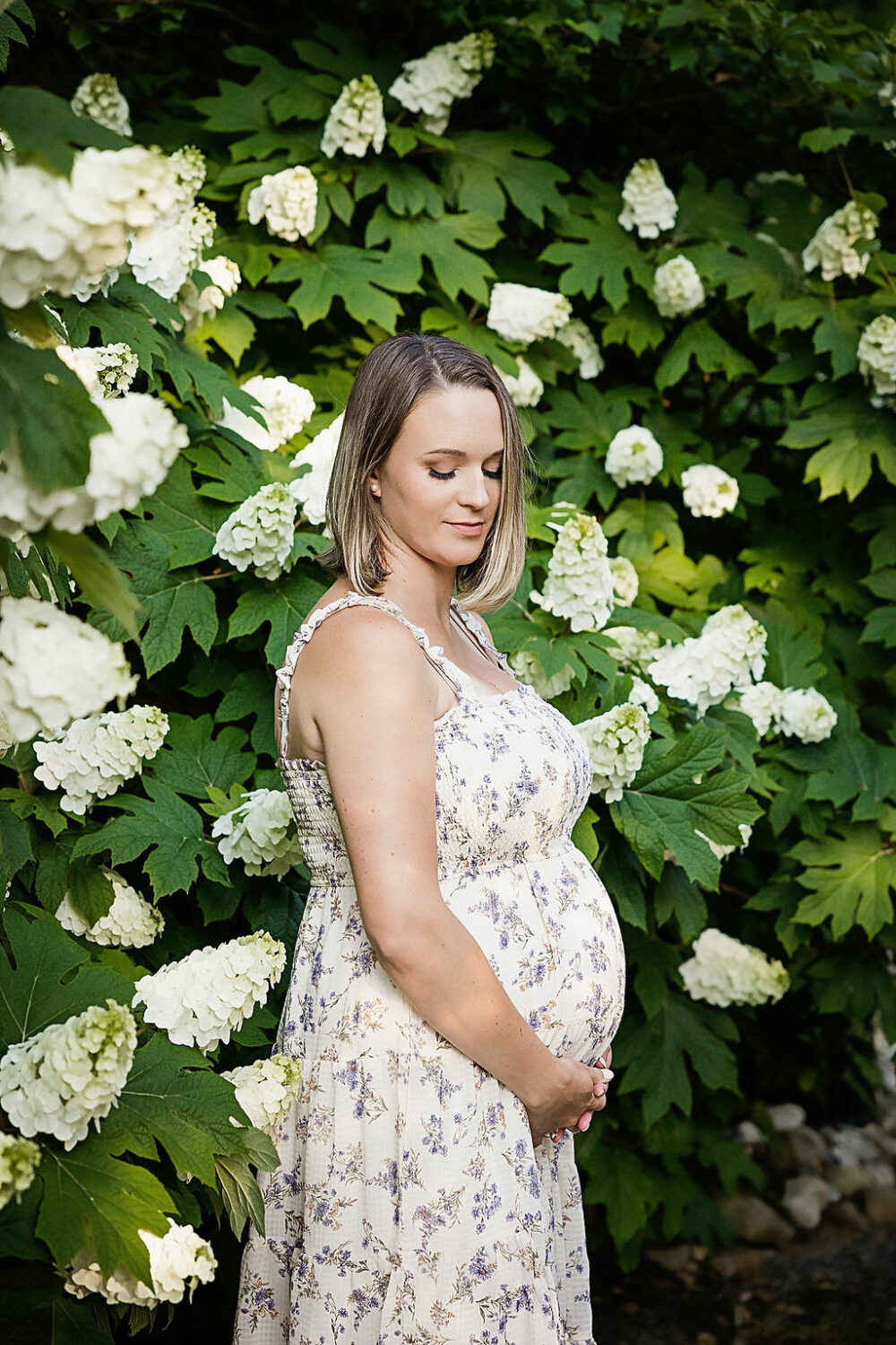 Mother to be wearing maternity gown holding her belly and looking down in front of white azalea flowers for her maternity portraits taken in Holmdel, New Jersey.