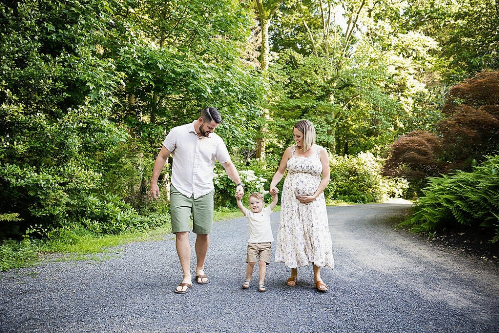 Mother and father holding toddler boy in between them holding hands while mother is holding her belly for their maternity session in Hamilton, New Jersey.