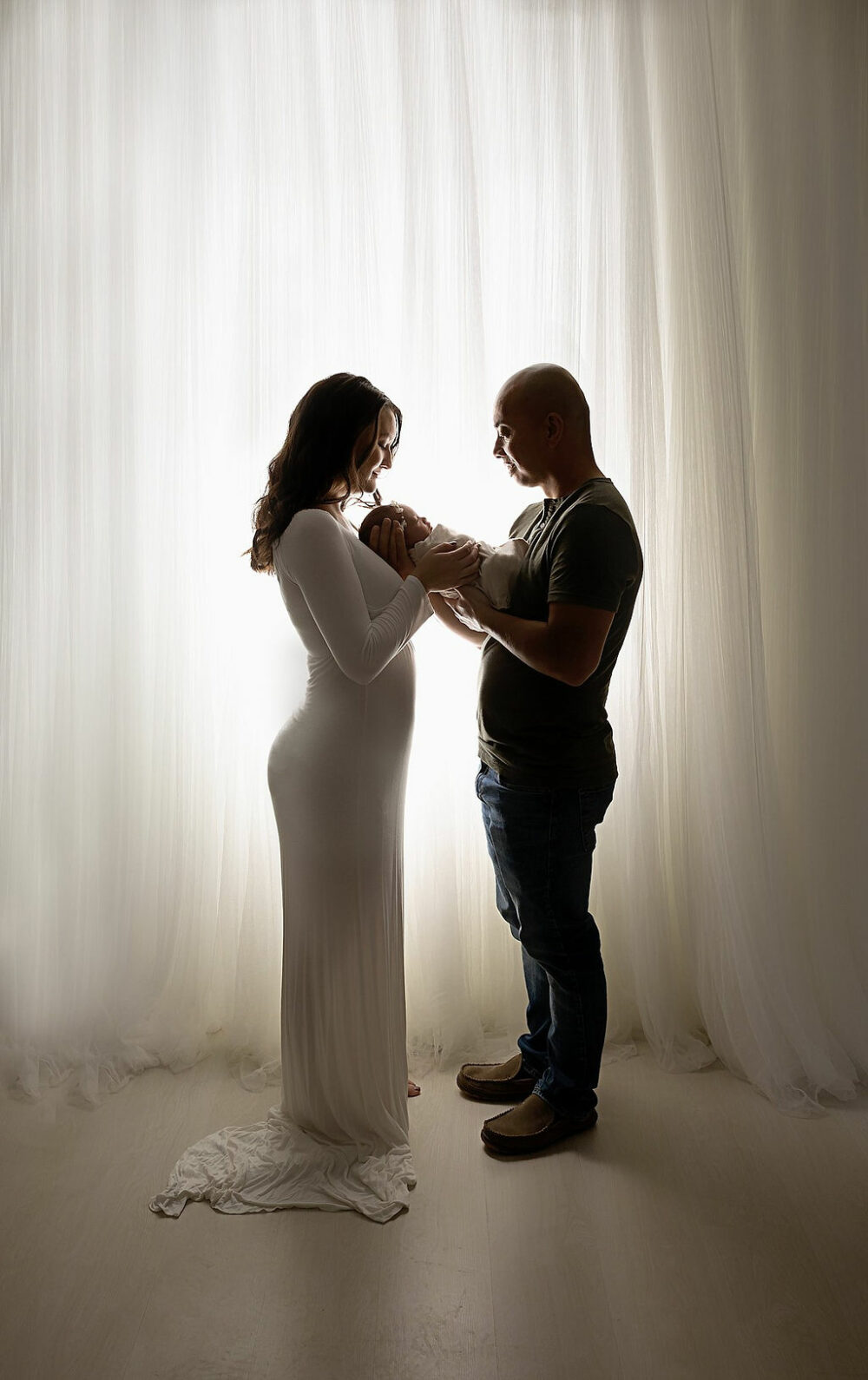 Light and bright photo of new parents holding baby girl between them wall facing each other looking at their newborn baby girl for their newborn milestones session in Mount Holly, New Jersey.