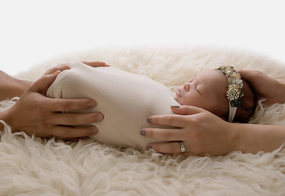 Close up of newborn girl sleeping on back wearing cream swaddle and matching headband with her parents hands holding her feet and shoulders on a textured blanket for her milestone newborn session taken in Pemberton, New Jersey.