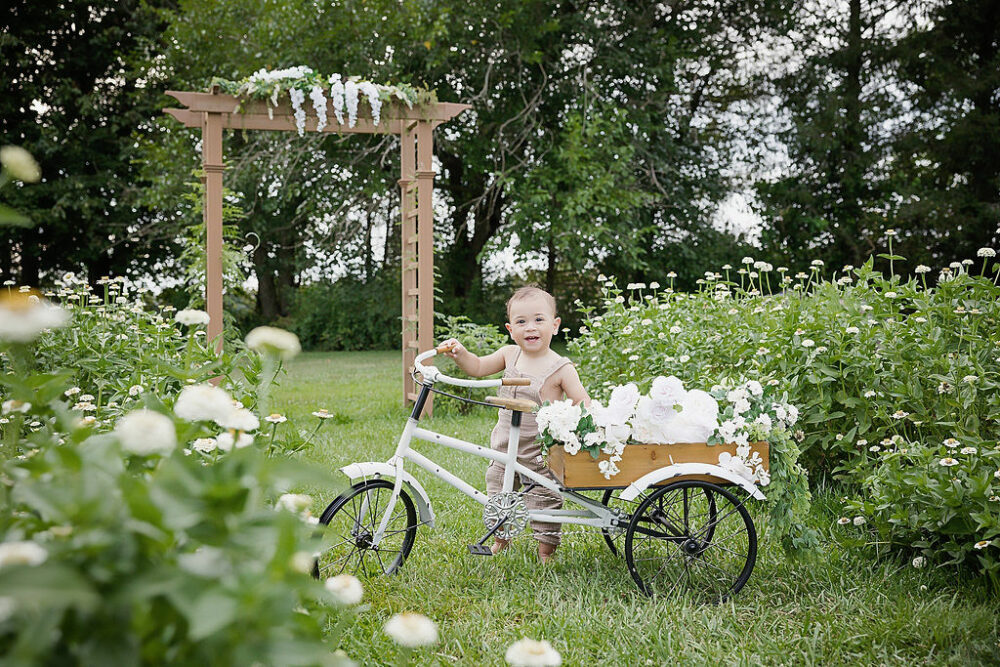 Toddler boy smiling a camera while holding onto photography bike prop and flower garden for flower mini sessions taken in Southampton, New Jersey.