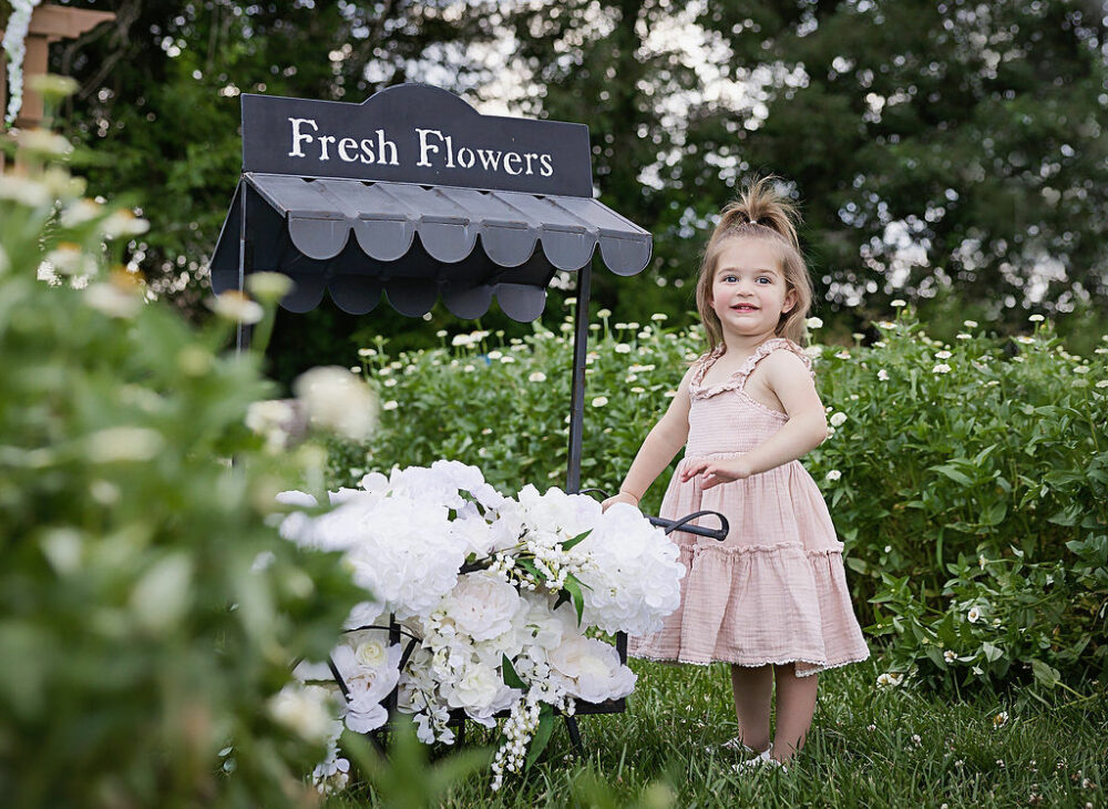 Toddler girl smiling at camera and Flowerfield holding prop for her summer pictures taken in Burlington, New Jersey.