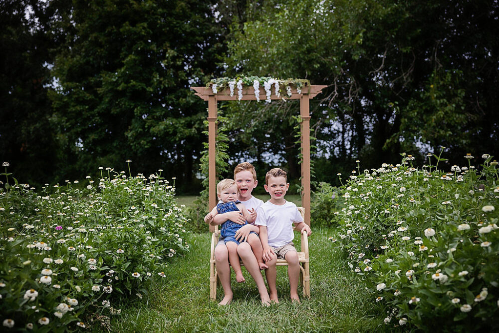 Three young brothers sitting on photography prop in middle of Flowerfield smiling at camera for their outdoor flower mini sessions taken in Hamilton, New Jersey.