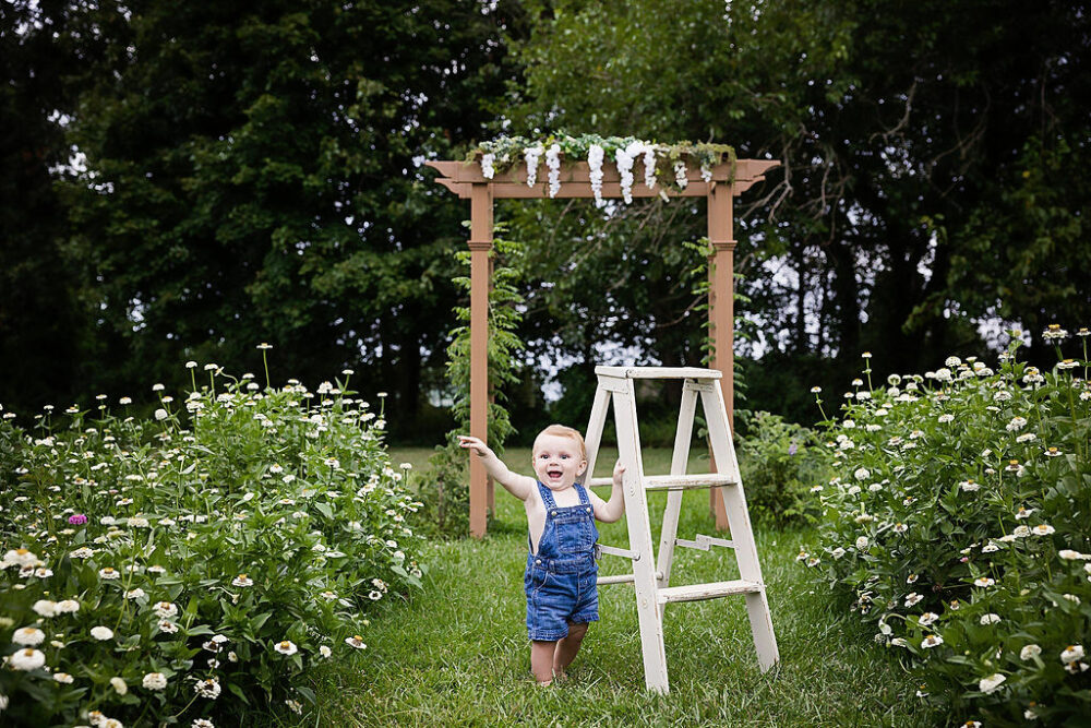Infant boy holding ladder photography prop in between Flowerfield smiling a camera for summer pictures taken in crosswicks, New Jersey.
