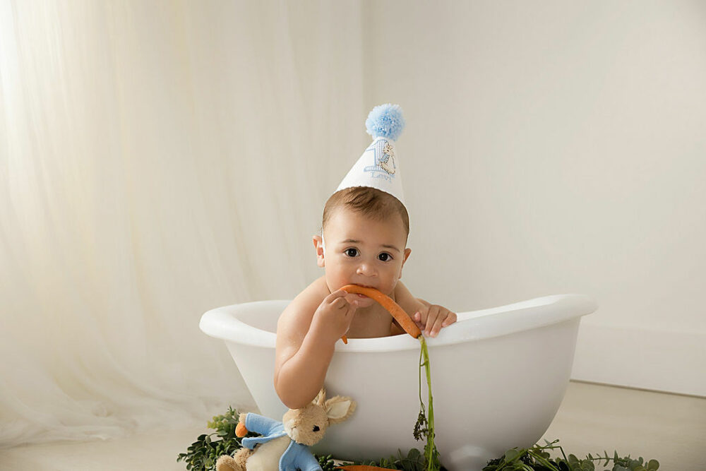 Toddler boy sitting in photography prop bath, tub, looking at camera and biting carrot and wearing birthday hat for his in studio Peter rabbit, first birthday session taken in Cherry Hill, New Jersey.